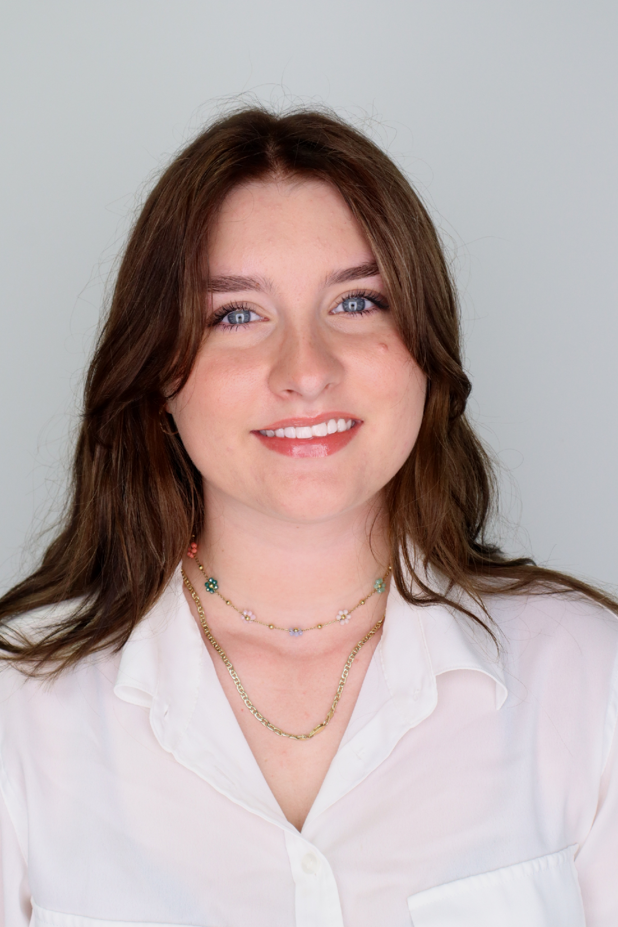 Natasha Bruce, Portrait. | Image description: She is wearing a white collared shirt, with a delicate necklace and her light brown hair parted down the middle. 